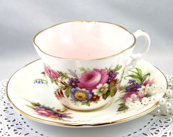 May include: A white teacup and saucer with a pink interior and a floral pattern in shades of pink, purple, blue, green, and yellow. The saucer has a gold rim.