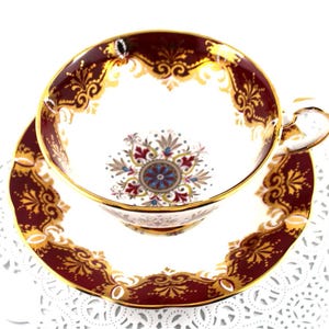 May include: A vintage teacup and saucer set with a burgundy and gold floral pattern. The cup and saucer have a white background with a gold rim.
