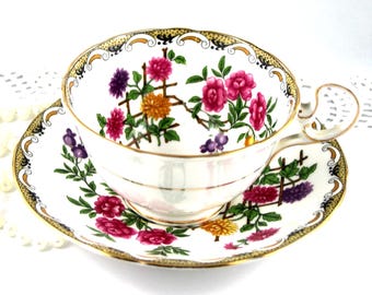 Könnte beinhalten: Eine weiße Teetasse und Untertasse mit einem Blumenmuster. Das Muster zeigt rosa, lila, gelbe und orangefarbene Blumen mit grünen Blättern auf weißem Hintergrund. Tasse und Untertasse haben einen goldenen Rand.