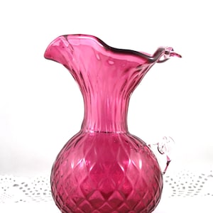 May include: A vintage cranberry glass pitcher with a ruffled top and a diamond-shaped pattern. The pitcher has a clear glass handle.
