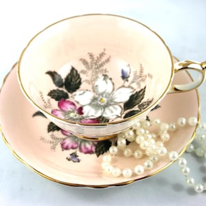Puede incluir: Un juego de té vintage rosa, compuesto por una taza y un platillo con un diseño floral de flores blancas y moradas, hojas verdes y ribetes dorados. Un collar de perlas descansa junto a la taza, realzando la estética elegante.