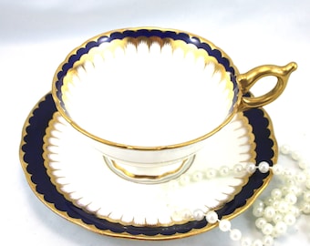 Può includere: Una tazzina da tè e un piattino bianchi con un bordo blu e dorato. Il piattino ha un bordo smerlato. La tazzina da tè ha un manico dorato. Una collana di perle bianche è in primo piano.