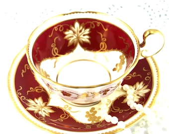 Può includere: Un set da tè in porcellana rossa e oro con un motivo a vite. Il piattino ha un bordo dorato e la tazza ha un manico dorato.