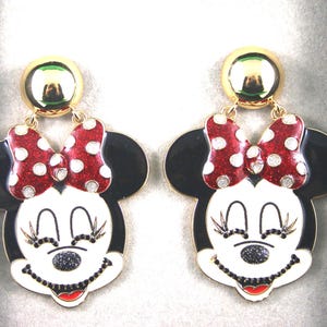 Op de afbeelding: Een paar gouden hangende oorbellen met een cartoon Minnie Mouse-ontwerp. De oorbellen hebben zwarte oren, een wit gezicht met zwarte ogen en een rode strik met witte stippen.