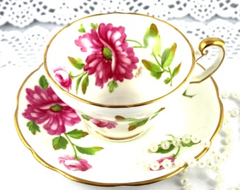 May include: A white teacup and saucer with a pink floral pattern and gold trim. The saucer has a scalloped edge. A string of pearls is draped over the saucer.