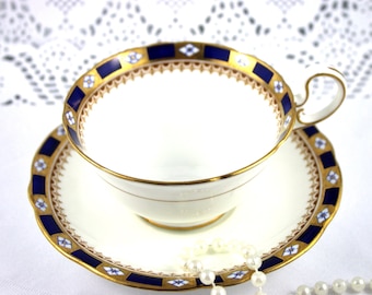 May include: A white teacup and saucer with a blue and gold trim. The saucer has a scalloped edge. A string of pearls is in front of the teacup and saucer.