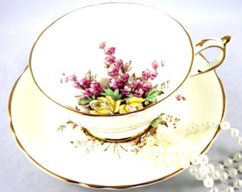 May include: A white teacup and saucer with a floral pattern featuring yellow roses and pink flowers. The saucer has a gold trim. A string of white pearls is lying on the saucer.