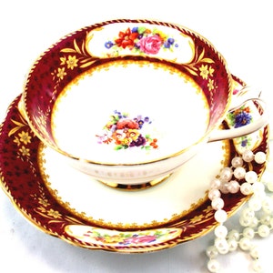 Op de afbeelding: Een vintage theekopje en schotelset met een bordeauxrood en gouden bloemenpatroon. Het kopje en de schotel hebben een witte achtergrond met een gouden rand en een bloemenontwerp in het midden. De schotel heeft een gegolfde rand.