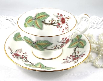 Könnte beinhalten: Eine weiße Teetasse und Untertasse mit einem floralen Muster in Grün-, Rot- und Goldtönen. Das Blumenmuster zeigt Blätter, Zweige und kleine Blüten.