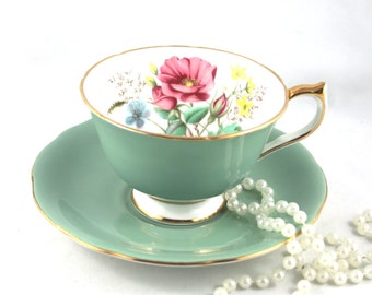 Könnte beinhalten: Ein Vintage-Teetassen- und Untertassen-Set in hellgrüner Farbe mit einem Blumendesign. Die Tasse hat einen goldenen Rand und die Untertasse einen goldenen Rand. Das Blumendesign zeigt eine rosa Rose, gelbe Blumen und grüne Blätter.