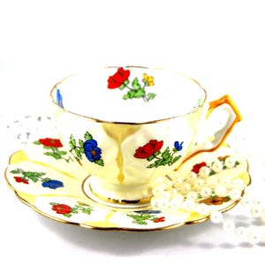 May include: A white porcelain teacup and saucer set featuring a delicate floral pattern with red, blue, and yellow flowers and green leaves. The rim of the cup and saucer is accented with gold trim, and the handle has a yellow and gold finish.