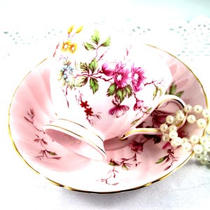 Könnte beinhalten: Eine rosa Teetasse und Untertasse mit floralem Design. Die Teetasse und Untertasse sind mit rosa, gelben und grünen Blumen und goldenen Verzierungen verziert. Eine Perlenkette liegt in der Nähe der Teetasse.