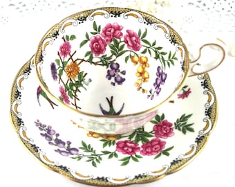 Könnte beinhalten: Eine weiße Teetasse und Untertasse mit einem Blumenmuster. Das Muster zeigt rosa, gelbe und violette Blumen sowie grüne Blätter und Vögel. Tasse und Untertasse haben einen goldenen Rand.