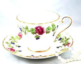 Könnte beinhalten: Eine weiße Teetasse und Untertasse mit einem floralen Muster in Rosa-, Grün- und Blautönen. Die Tasse hat einen goldenen Rand und einen zarten Henkel. Die Untertasse hat ein passendes Blumenmuster und einen goldenen Rand.