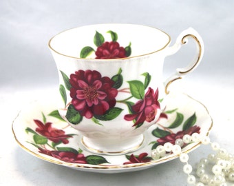 May include: A white teacup and saucer with a floral design featuring red and green flowers. The cup and saucer have a gold trim.