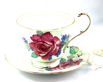 May include: A vintage teacup and saucer set with a green background and a large pink rose with green leaves and blue flowers. The set has a gold trim.