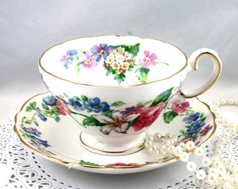 Può includere: Una tazza da tè e un piattino bianchi con un motivo floreale in tonalità di rosa, blu e verde. La tazza e il piattino hanno un bordo dorato.