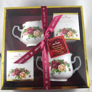 Könnte beinhalten: Ein Geschenkeset mit Royal Albert Tee-Accessoires, bestehend aus zwei Teetassen mit Blumenmustern, einer Schachtel Earl Grey Teebeutel und einer Schachtel English Afternoon Teebeutel. Das Set ist mit einem roten Band gebunden und kommt in einer goldenen und roten Schachtel.