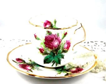 May include: A white teacup and saucer set with a gold rim and a floral rose design. The roses are a deep pink with green leaves. A pearl necklace is draped near the saucer.