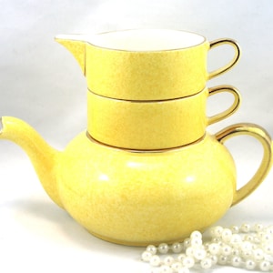 Puede incluir: Una tetera amarilla con un borde y un asa dorados, apilada con dos tazas amarillas con bordes y asas dorados. Las tazas están anidadas dentro de la tetera. Una cadena de perlas blancas está en primer plano.