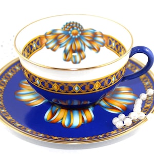 May include: A blue and white teacup and saucer set with a floral design. The cup and saucer have gold trim and a blue handle. The floral design features orange and blue ribbons. A string of pearls is next to the saucer.