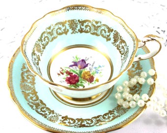 May include: A vintage teacup and saucer set in a light blue hue, adorned with gold filigree and trim. The teacup features a floral bouquet in the center. A pearl necklace rests beside the teacup and saucer.