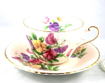 May include: A white teacup and saucer set with a floral design. The cup and saucer are decorated with yellow, purple, and pink flowers and green leaves. The rims are trimmed with gold. A classic tea set.
