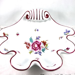 May include: White ceramic serving dish with a scalloped edge and a floral design. The dish has a deep burgundy trim and a central arrangement of pink and purple flowers with green leaves. The dish has a decorative handle.