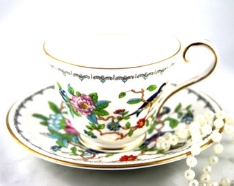 Könnte beinhalten: Eine weiße Teetasse und Untertasse mit einem floralen und Vogel-Design. Tasse und Untertasse haben einen goldenen Rand und ein zartes Blumenmuster mit blauen, rosa und grünen Akzenten. Die Untertasse hat ein passendes Blumenmuster.