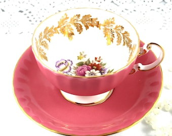 May include: A pink teacup and saucer set with a floral design and gold trim. The cup has a white interior with a gold leaf design around the rim. The saucer has a scalloped edge.