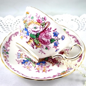 Puede incluir: Juego de taza y platillo de té blanco con diseño floral. La taza y el platillo están decorados con flores rosas, azules, amarillas y verdes. Los bordes están adornados con dorado. Un collar de perlas está cerca de la taza.
