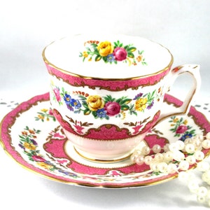 Könnte beinhalten: Eine rosa und weiße Teetasse und Untertasse mit einem Blumenmuster und goldenen Verzierungen. Die Untertasse hat eine Perlenkette darauf.
