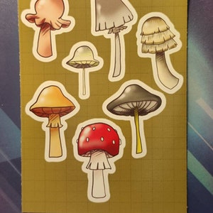 May include: A collection of nine colourful mushroom stickers with various shapes and sizes. The stickers feature a variety of colours, including red, orange, yellow, white, and grey.