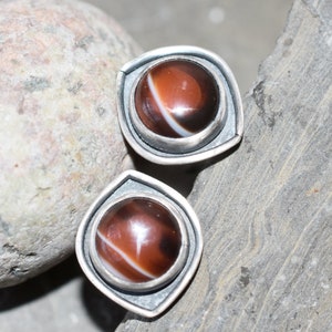 May include: A pair of stud earrings featuring a polished, round, brown stone with a white stripe. The stones are set in a silver-colored metal with a square, geometric design.
