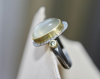 May include: A silver ring with a large oval-shaped moonstone set in a gold bezel. The ring is accented with small diamonds and a green gemstone.