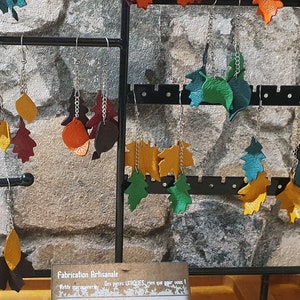 Op de afbeelding: Een display met leren oorringen in de vorm van bladeren in verschillende kleuren, waaronder rood, oranje, geel, groen en bruin. De oorringen worden gepresenteerd op een zwarte metalen standaard met haken. Het bord eronder vermeldt "Fabrication Artisanale" en "Le cuir".