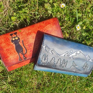Peut inclure: Deux portefeuilles en cuir sur l'herbe. L'un est orange avec une illustration de chat noir. L'autre est bleu avec un motif de montagne et de forêt. Les deux sont rectangulaires avec un rabat.