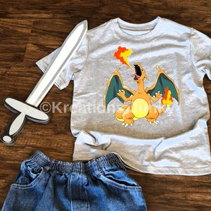 May include: A gray t-shirt with a graphic of a cartoon orange dragon breathing fire. The dragon is on a gray t-shirt with a white collar. The t-shirt is laying on a wooden surface with a toy sword.