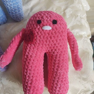 Może przedstawiać: Różowa pluszowa zabawka wykonana na szydełku z czarnymi oczami i białym otworem gęby. Zabawka ma dwie ręce i dwie nogi.