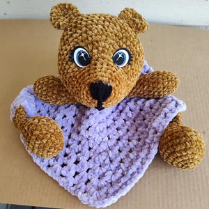 May include: A brown crocheted teddy bear with sparkly blue eyes is holding a lavender crocheted blanket.