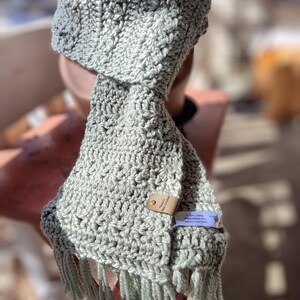May include: A sage green crocheted scarf with fringe. The scarf has a leather tag with the text "Handmade with Love" on it.