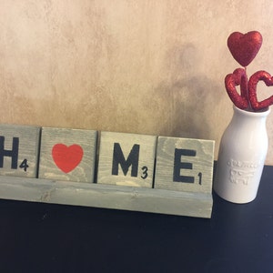 May include: Wooden block letters spelling "HOME" with a red heart in place of the "O". The letters are on a wooden stand. A white vase with three red glitter hearts is on the right side of the image.