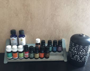 May include: A set of 17 essential oil bottles in various colors, arranged on a wooden shelf with a black metal diffuser in the background.