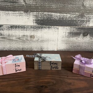 May include: Three stacked wooden boxes with pink, blue, and purple paint. The boxes are tied with pink and white gingham ribbon. The top box of each stack has a different phrase written on it: "Tea Time", "Farm Fresh", and "Fresh Lavender".