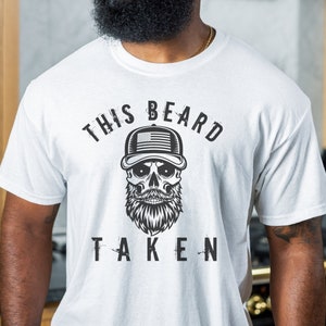 May include: White t-shirt with a black skull wearing a baseball cap with an American flag design. The text "This Beard Taken" is printed on the shirt.