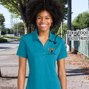 May include: Teal polo shirt with a three-button placket and a small logo on the left chest. The logo features the letters "CAG" above the text "C.A. GRAY JUNIOR HIGH SCHOOL". The shirt is worn by a person smiling in front of a street.