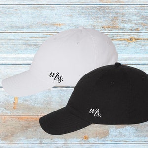 K&ouml;nnte beinhalten: Zwei Baseballkappen, eine wei&szlig;e und eine schwarze, werden gezeigt. Auf der wei&szlig;en Kappe ist "Mrs." in schwarzer Schrift auf der Seite gestickt, w&auml;hrend die schwarze Kappe "Mr." in wei&szlig;er Schrift hat. Die Kappen befinden sich auf einer hellblauen Holzoberfl&auml;che.
