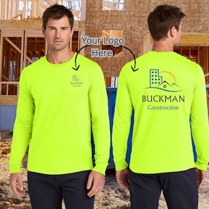 May include: Neon yellow long-sleeve shirt with the "BUCKMAN Construction" logo on the front and back. The logo features a building, sun, and mountain design. The shirt is worn by a person.