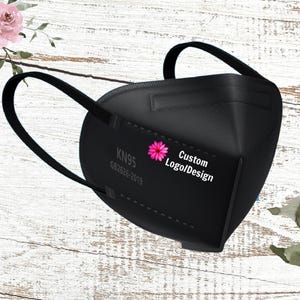 May include: Black KN95 face mask with ear loops. The mask has the text "KN95 GB2626-2019" and "Custom Logo/Design" with a pink flower graphic. The mask is set against a white wood grain background with floral accents.