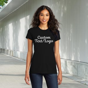 May include: A black t-shirt with the text "Custom Text/Logo" in white script. The shirt is short-sleeved and appears to be made of a soft material. The person is wearing blue jeans.